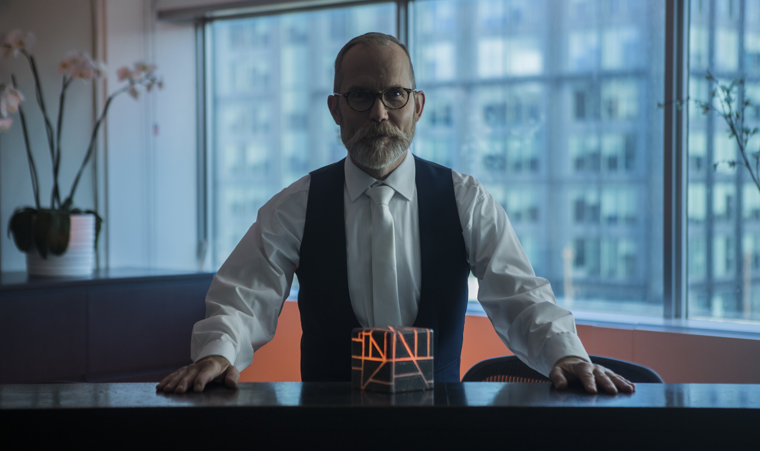 Still from Transference, a man in a waistcoat standing at a table in a glass office