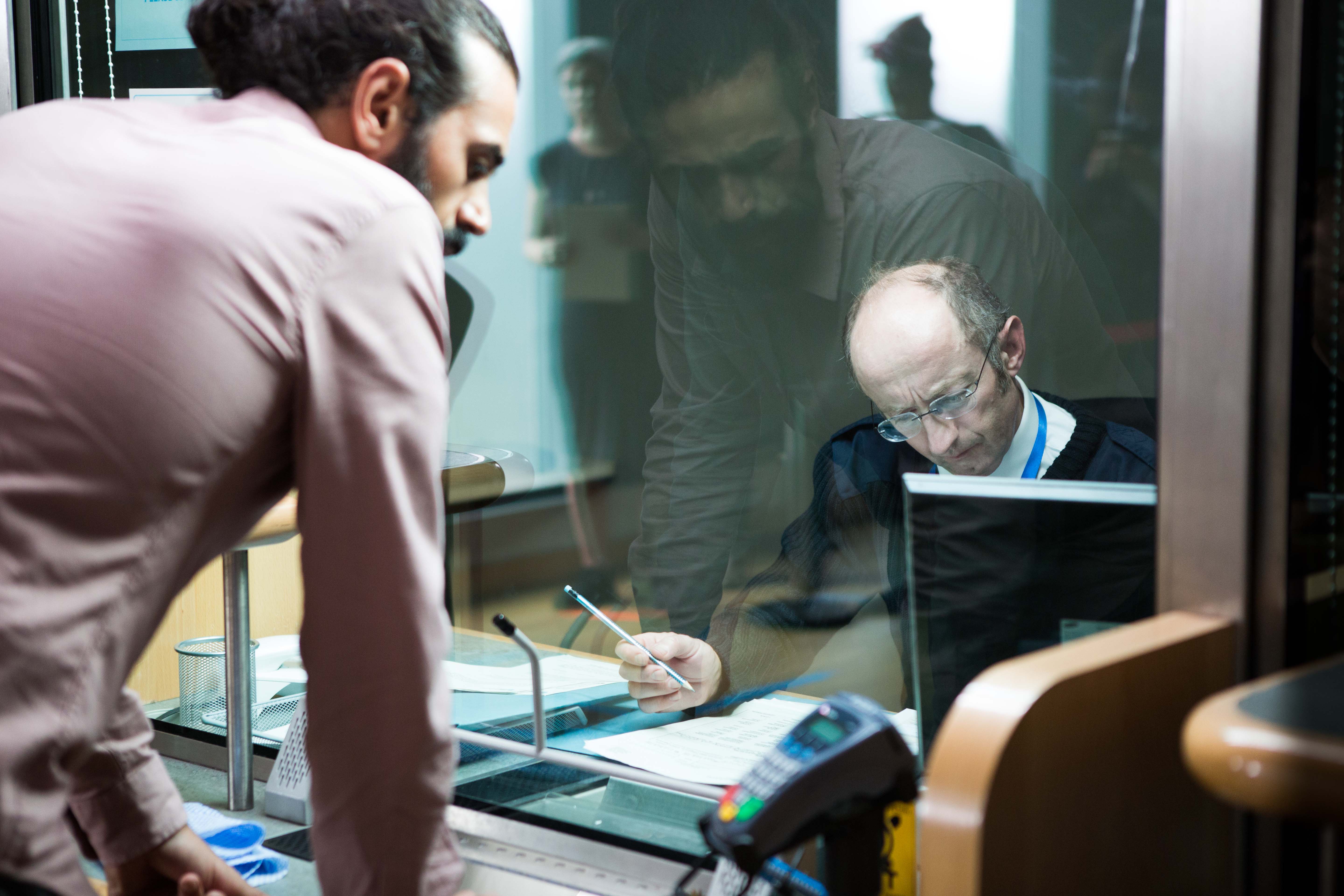 Still from Leave to Remain, a man leaning over a counter as a clerk writes