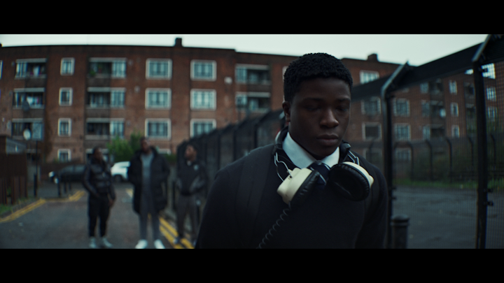 Still from 1 UP, the lead walking through the estate in school uniform with headphones round his neck